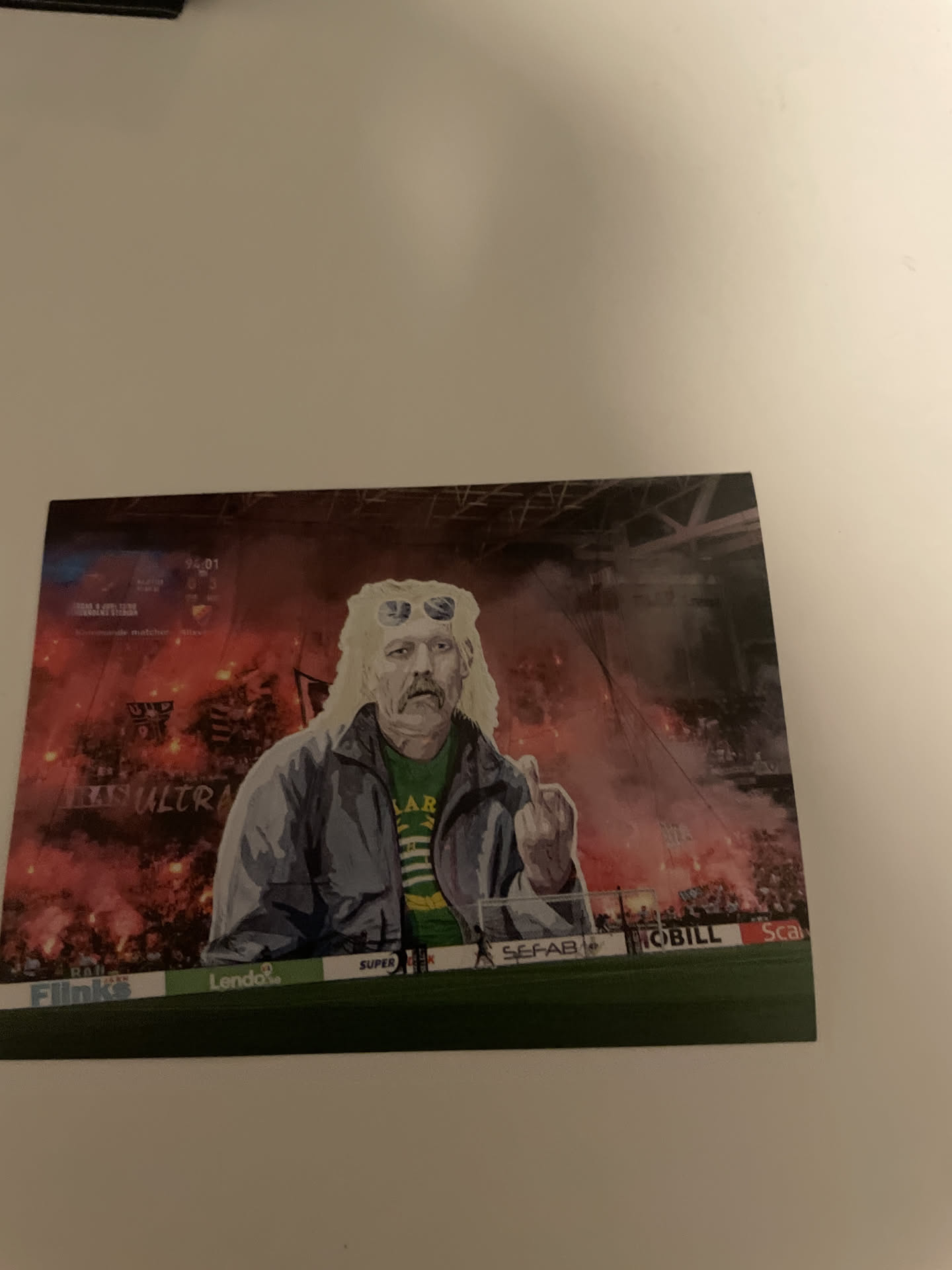 The sticker features a stylized illustration of a man with long, wavy blonde hair, a prominent mustache, and white sunglasses pushed up onto his head. He is depicted wearing a grey jacket over a green and yellow striped t-shirt, with the letters 'LAR' visible on it. His right hand is raised, making a middle finger gesture. The background is a dynamic scene of a stadium at night, illuminated by numerous red flares and smoke, suggesting an intense football match or event. A scoreboard in the upper left shows '94:01' and some obscured scores, with the word 'ULTRA' partially visible below it. Along the bottom, a green barrier displays various sponsor logos including 'Flinks', 'Lendo', 'SUPER', 'SEFAB', 'MOBILL', and 'Scal'. The overall aesthetic is gritty and energetic, with a dominant color palette of reds, oranges, and dark tones contrasting with the figure's lighter features.