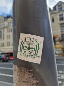 A sticker featuring the coat of arms of Hamar, Norway, with the year 1918 prominently displayed. The design is simple, using a cream background with green and black detailing. The sticker is affixed to a grey pole in an urban setting.