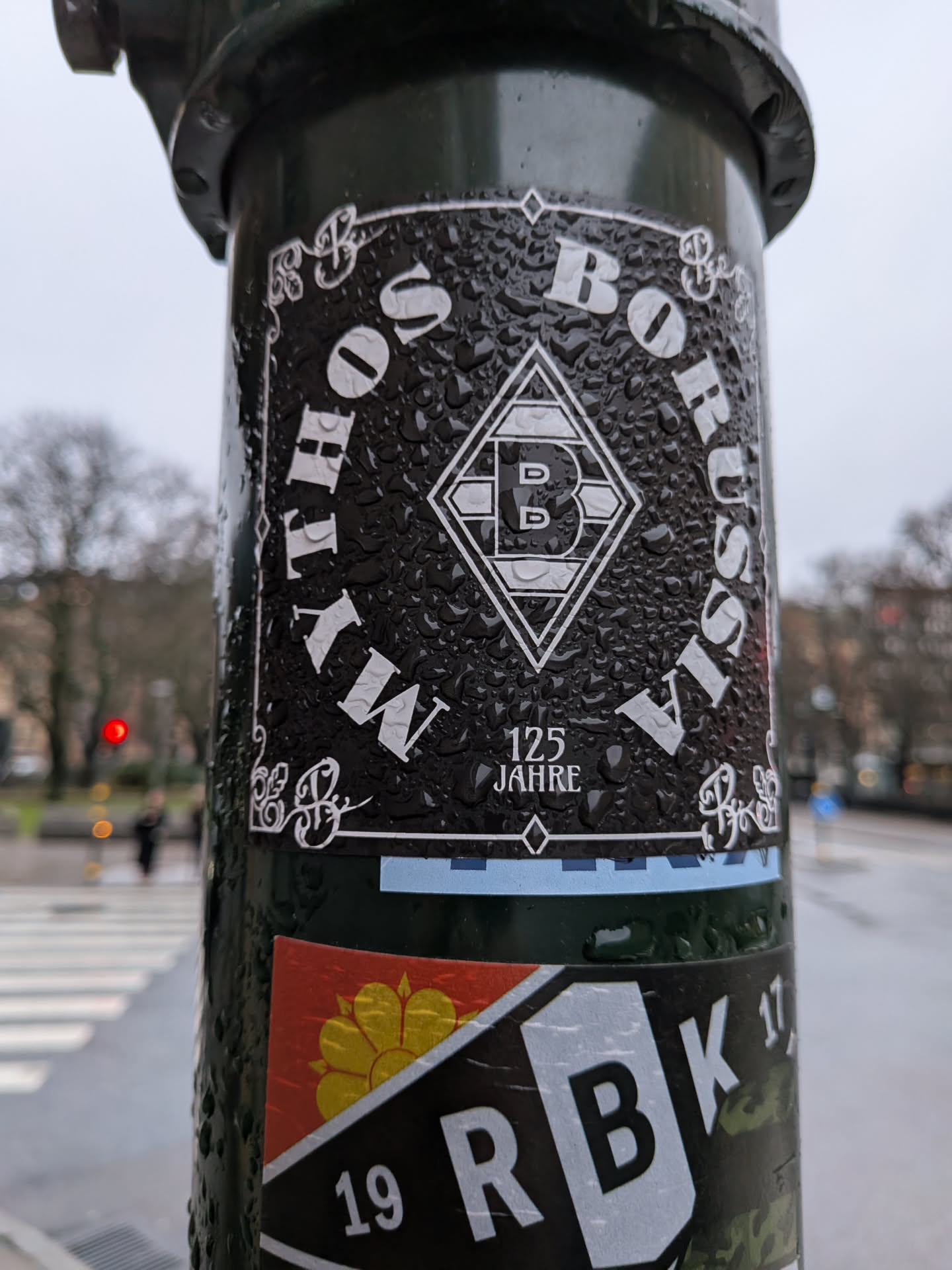 The sticker is predominantly black with white lettering and graphics. It features the text 'MY BORUSSIA' in a circular arrangement around a diamond-shaped logo. The logo contains the letters 'BMG' stylized within the diamond. Below the logo, it reads '125 JAHRE' (125 Years). The sticker has a textured appearance due to numerous water droplets on its surface. Decorative scrollwork is present in the corners. A thin blue stripe runs along the bottom edge of this sticker, and another sticker with a red and yellow emblem and the text '19 RBK 17' is partially visible below it.