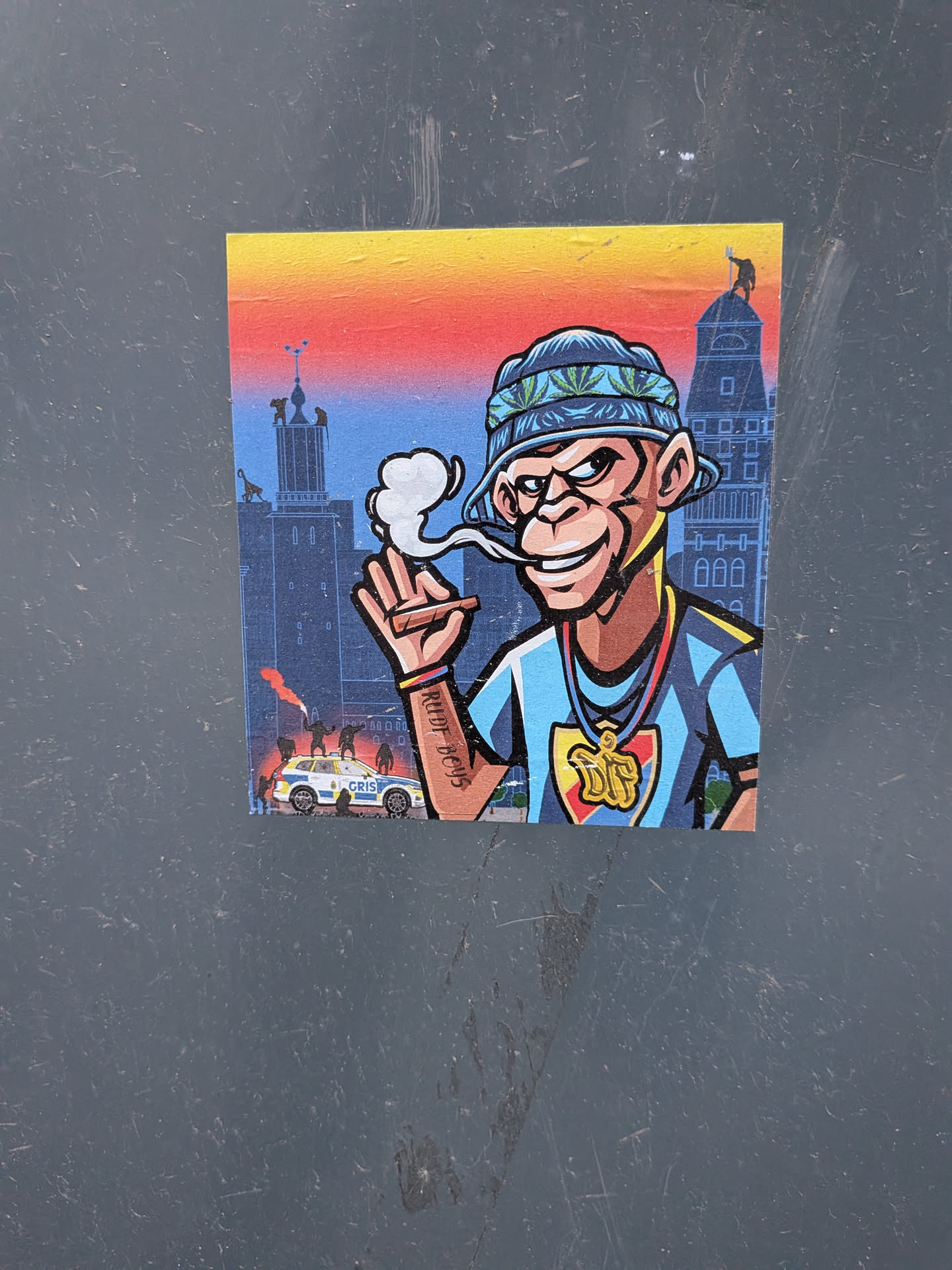 The sticker features a cartoon-style monkey character with brown fur, a wide grin, and a mischievous look. It wears a blue bucket hat adorned with green cannabis leaves and a light blue t-shirt with dark blue stripes. Around its neck is a gold chain with a large 'DIF' pendant in gold, red, and yellow. The monkey is holding a lit cigar or joint, from which a puff of white smoke emanates. On its right forearm, a tattoo reads 'RUDE BOYS'. The background depicts a stylized urban cityscape with tall buildings under a gradient sky transitioning from yellow to orange to red and then blue. In the lower left, a white and blue police car with 'GRIS' written on its side is visible, with small figures and a red flare in the foreground.