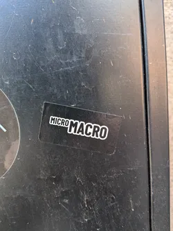 Street sticker A rectangular black sticker featuring the words 'MICRO MACRO' in bold, white, sans-serif lettering with a white outline. The word 'MICRO' is smaller and positioned slightly above the baseline of the larger, more prominent 'MACRO'. The sticker has a clean, high-contrast, minimalist design and appears to be applied to a dark, scratched surface.