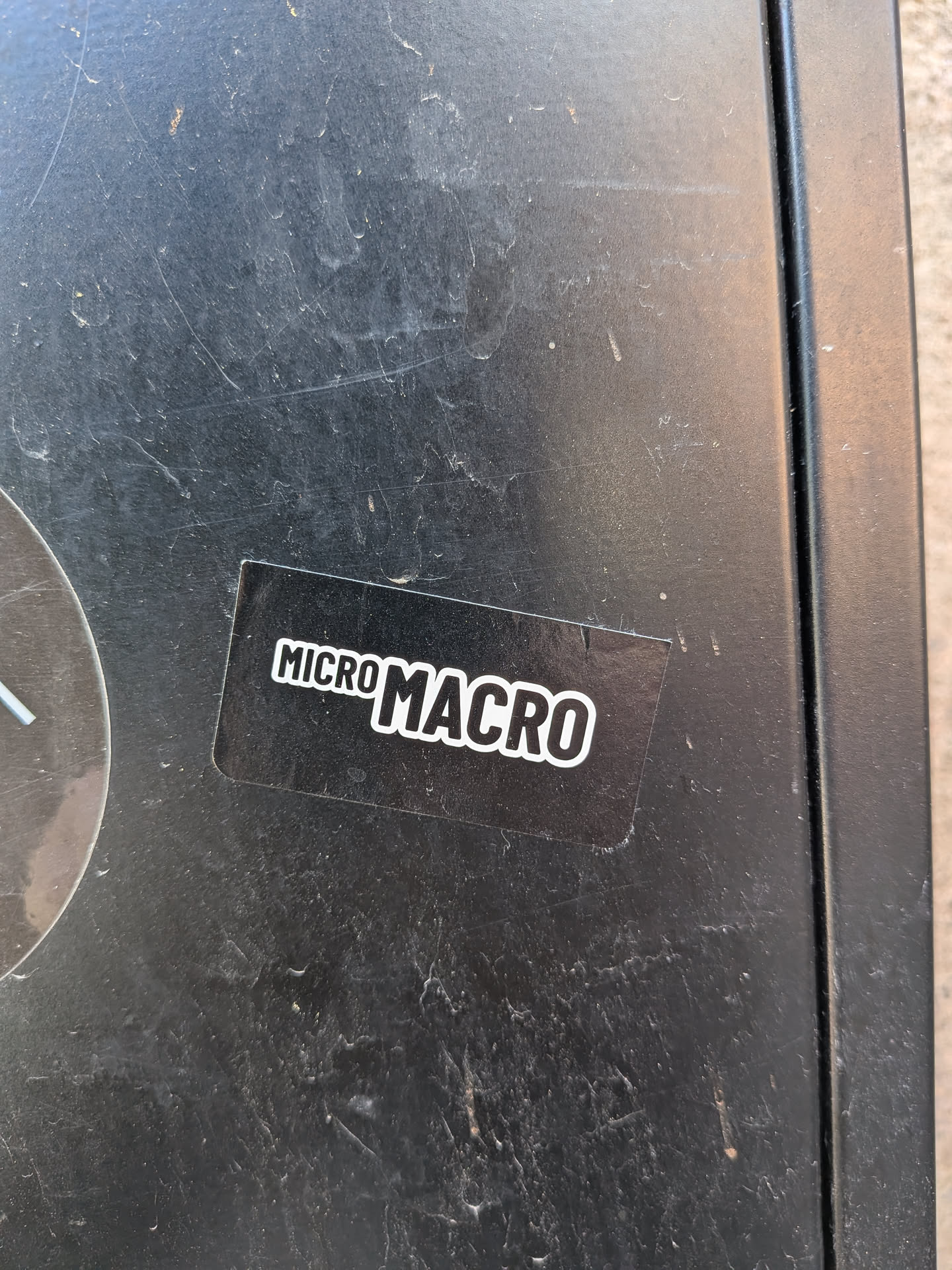 A rectangular black sticker featuring the words 'MICRO MACRO' in bold, white, sans-serif lettering with a white outline. The word 'MICRO' is smaller and positioned slightly above the baseline of the larger, more prominent 'MACRO'. The sticker has a clean, high-contrast, minimalist design and appears to be applied to a dark, scratched surface.