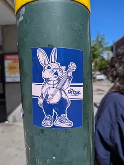 Street sticker The sticker features a stylized, anthropomorphic rabbit character rendered in white outline against a solid dark blue background. The rabbit is standing upright, wearing what appears to be a jacket or shirt and pants, and sneakers. It has large ears, whiskers, and an open mouth, suggesting it's singing or calling out. The rabbit is holding and playing a banjo. A white horizontal stripe runs across the middle of the sticker, behind the rabbit, containing the text "THE PATZAC ILLUSTRATOR" in black. The overall style is illustrative and cartoonish, with clear lines and a two-tone color scheme of dark blue and white.