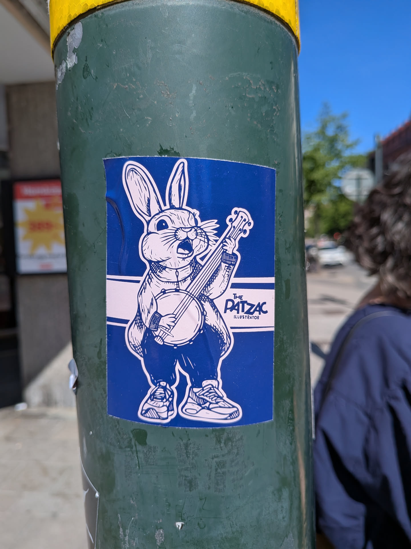 The sticker features a stylized, anthropomorphic rabbit character rendered in white outline against a solid dark blue background. The rabbit is standing upright, wearing what appears to be a jacket or shirt and pants, and sneakers. It has large ears, whiskers, and an open mouth, suggesting it's singing or calling out. The rabbit is holding and playing a banjo. A white horizontal stripe runs across the middle of the sticker, behind the rabbit, containing the text "THE PATZAC ILLUSTRATOR" in black. The overall style is illustrative and cartoonish, with clear lines and a two-tone color scheme of dark blue and white.