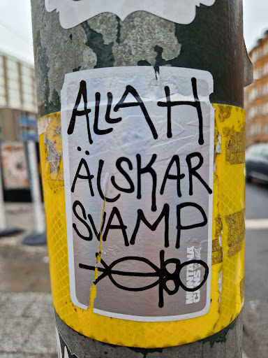 A sticker featuring the text "Allah Älskar Svamp" (God loves mushrooms) in black marker on a silver background. There is a simple drawing of a mushroom at the bottom. The sticker is slightly torn. The sticker is on a yellow reflective band around a pole.