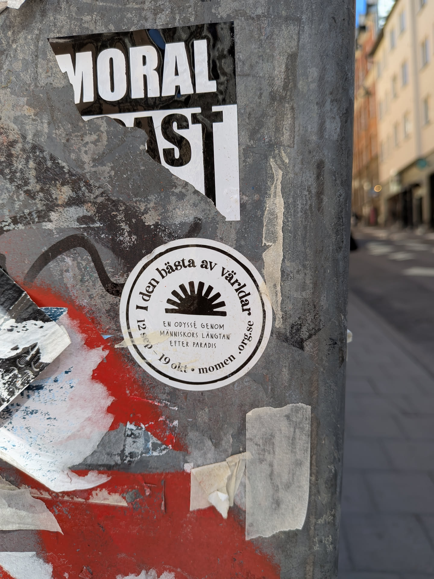 A circular white sticker with black text and a central graphic, affixed to a weathered metal pole. The sticker features a stylized black sun or rising sun symbol with radiating rays at its center. Around the outer edge, text is arranged in a circle. Below the sun symbol, there are two lines of text. The sticker shows some minor wear and tear, consistent with being outdoors.
