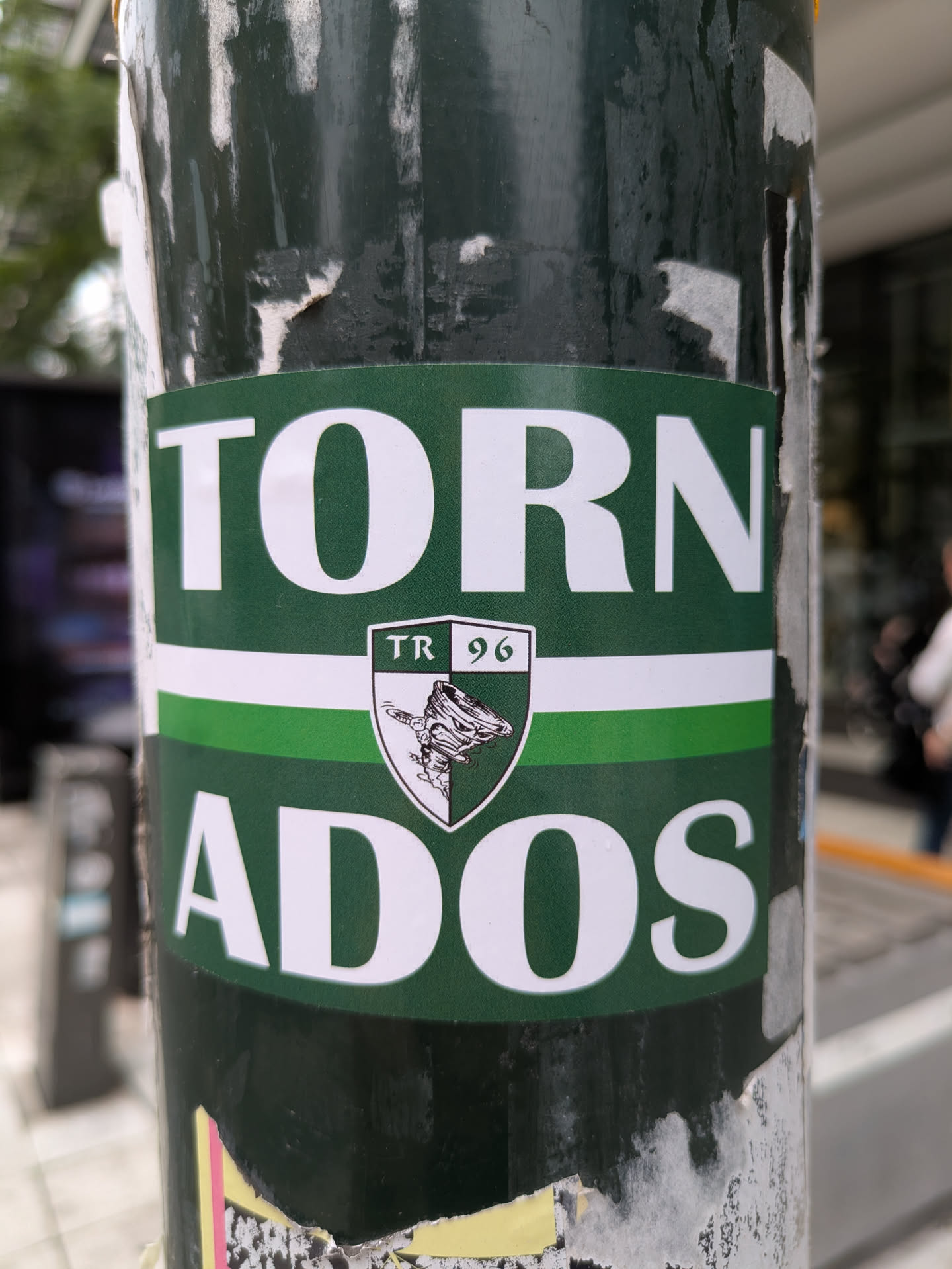The sticker is rectangular with a dark green background. The word "TORN" is displayed in large white capital letters at the top, and "ADOS" is similarly displayed at the bottom, together forming "TORNADOS". A white horizontal stripe separates the two words, with a lighter green stripe beneath it. Centered on these stripes is a shield-shaped emblem. The top section of the shield reads "TR 96". The bottom section of the shield features a graphic of a tornado. The shield itself is split vertically with dark green and white sections. The sticker is applied to a dark green pole and shows signs of wear and peeling.