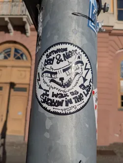 Street sticker The sticker is circular with a jagged, serrated edge. It features a black and white illustration of a stylized wolf's face with sharp teeth, wide eyes, and a crown-like element on its head. Lightning bolts are incorporated into the design. Text is present in a distressed, handwritten style, arranged in arcs around the wolf's face. The background is white, and the overall aesthetic is bold and graphic.