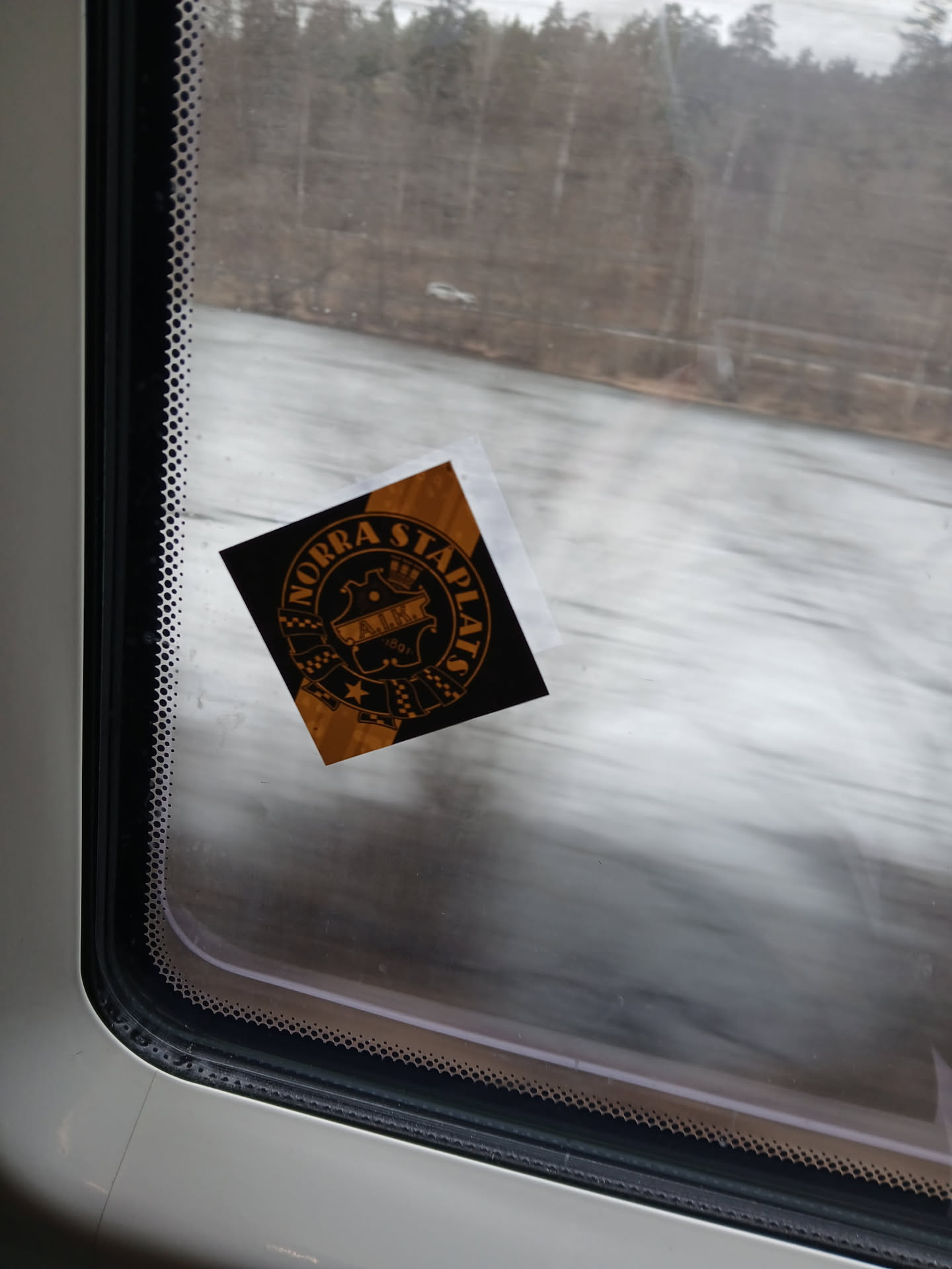 A square-shaped sticker, predominantly black with metallic gold or orange details, featuring a circular crest design. The crest includes a central shield-like emblem topped with a crown. Inside the shield, the letters 'A.I.K.' are prominently displayed, with '1891' beneath them. The text 'NORRA STAPLATS' is arched along the top inner edge of the circular border. The design also incorporates checkered patterns on the sides of the central shield and a star at the bottom. The overall aesthetic is heraldic and vintage, with a slight corner of the sticker appearing to peel from the window surface.