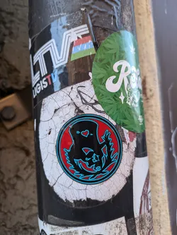 Street sticker A round sticker featuring a cartoon creature with a top hat and small hearts for eyes, surrounded by stylized foliage. The sticker has a red background with a teal outline and is applied to a weathered surface.