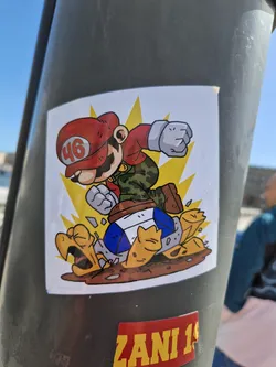 Street sticker This sticker shows a stylized character resembling Mario from the Super Mario series, wearing a red cap with the number 46, kicking a Koopa Troopa. The art style is cartoonish and vibrant. The background is a simple yellow burst. The sticker is affixed to a gray pole.