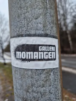 A black and white sticker on a metal pole, possibly advertising an art gallery named Momangen.
