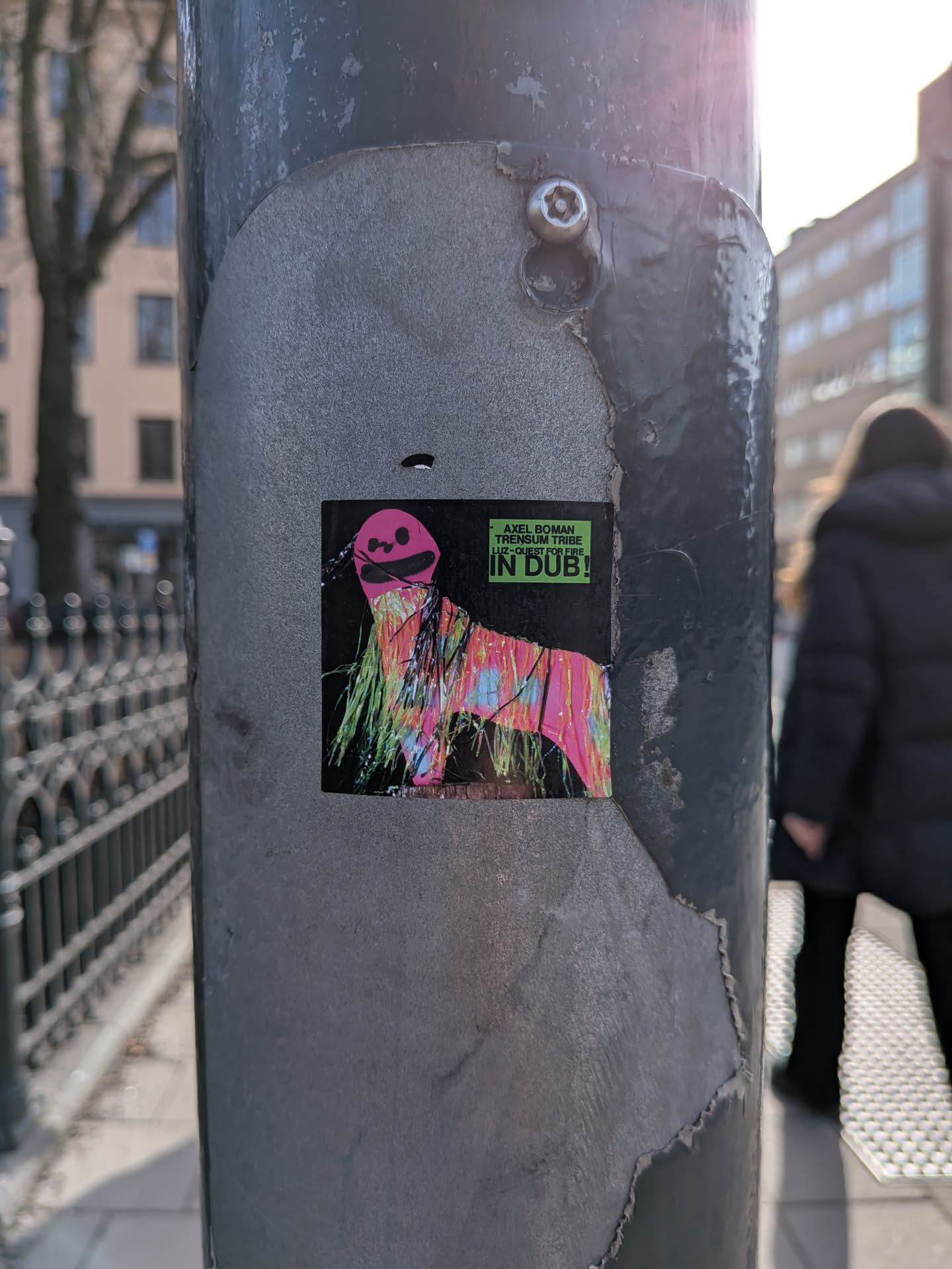 The sticker features a stylized, abstract creature against a solid black background. The creature has a bright pink, smiling, somewhat mask-like head with two black dots for eyes. Its body is composed of numerous vertical, shaggy strands or 'fur' in a vibrant, psychedelic mix of neon green, bright pink, light blue, and yellow, giving it a dynamic, almost dripping appearance. The overall form suggests a four-legged animal or a person in a shaggy costume. In the top right corner, there's a small rectangular green box with white text.