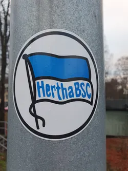 Street sticker A round sticker featuring a blue flag with the text "HerthaBSC" on it. The sticker is affixed to a gray metal pole.