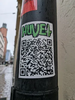 Street sticker A rectangular sticker with a white background, prominently featuring the word 'HUVE+' at the top in a vibrant green, graffiti-style font with a black outline. Below the text, a large, black and white QR code occupies the majority of the sticker's surface. The sticker is affixed to a dark, possibly black, utility pole and shows signs of outdoor exposure, including visible water droplets.
