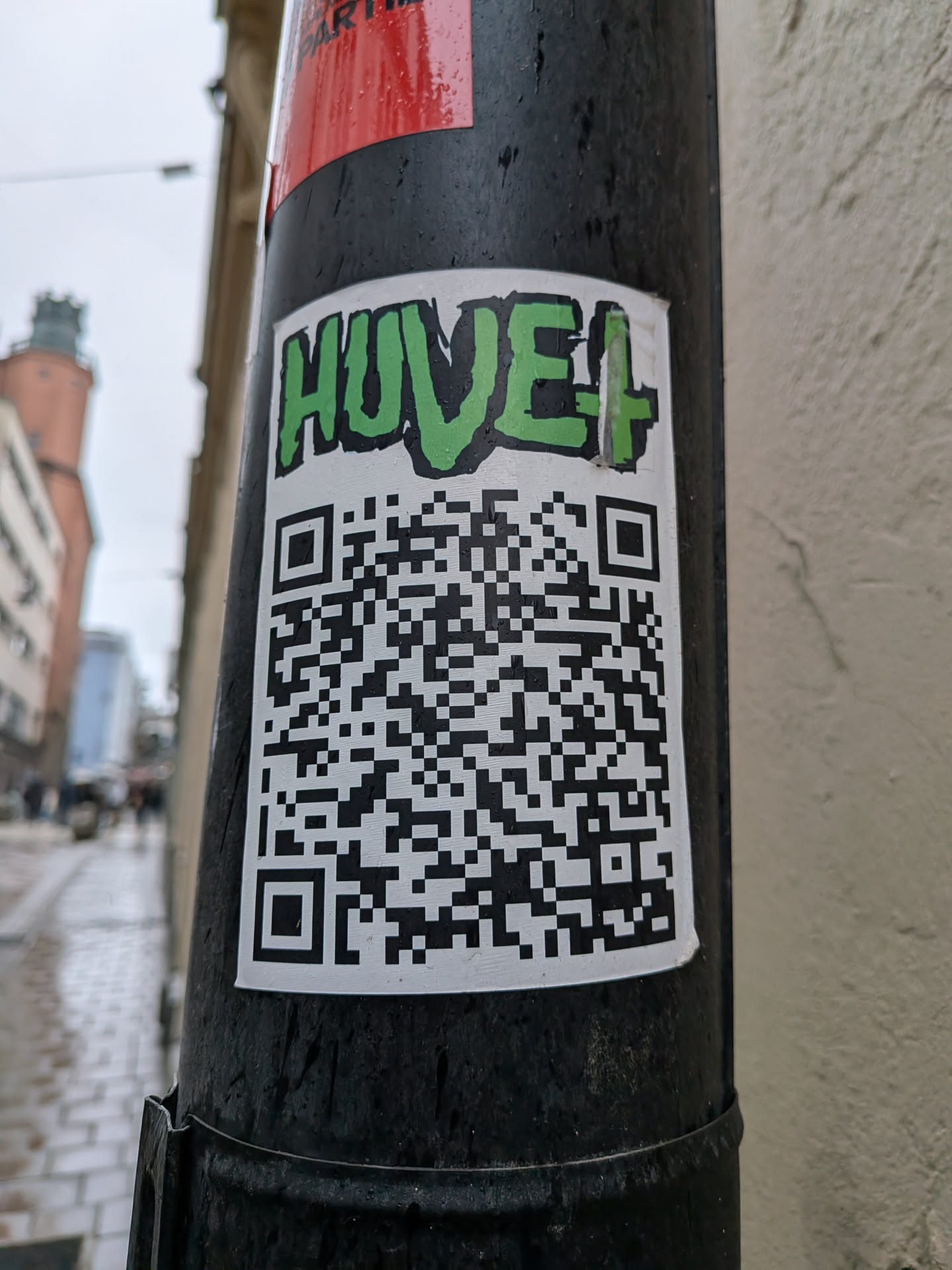 A rectangular sticker with a white background, prominently featuring the word 'HUVE+' at the top in a vibrant green, graffiti-style font with a black outline. Below the text, a large, black and white QR code occupies the majority of the sticker's surface. The sticker is affixed to a dark, possibly black, utility pole and shows signs of outdoor exposure, including visible water droplets.