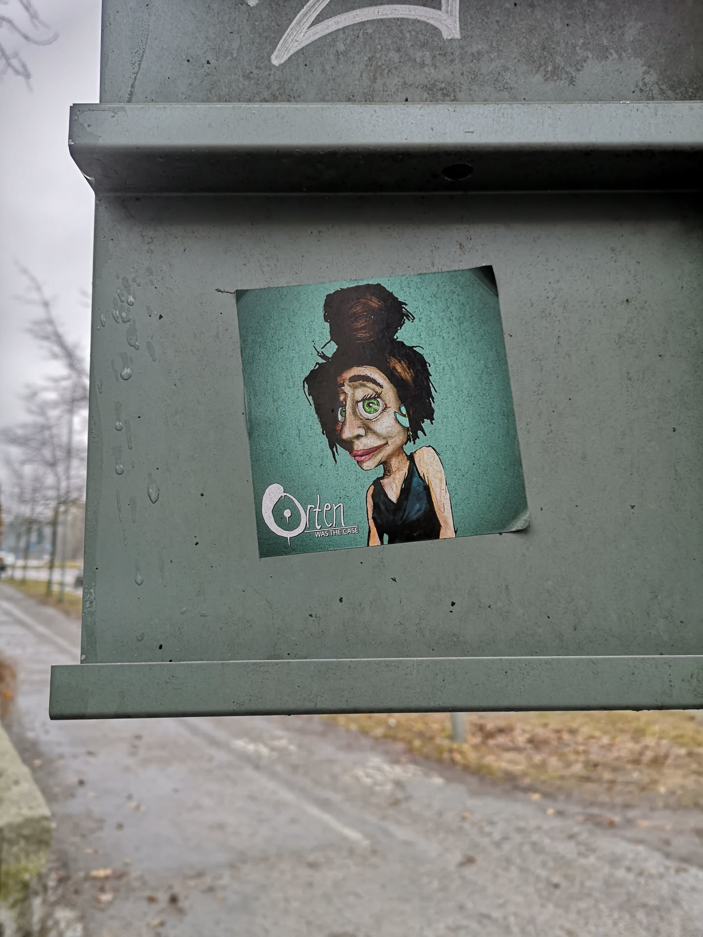 The sticker features a stylized, cartoonish portrait of a woman from the chest up, set against a gradient background that transitions from a darker teal at the top to a lighter, almost mint green at the bottom. The woman has dark, messy hair styled in a large, high bun. Her face is elongated with prominent features: large, bright green eyes, thick eyebrows, a slightly upturned nose, and full, pink lips. She has a pale complexion with subtle hints of red on her cheeks and nose. She wears a dark, sleeveless top. A small, light blue or teal earring is visible on her right ear. The art style is illustrative and somewhat whimsical, conveying a thoughtful or slightly melancholic expression. In the bottom left corner, white text reads 'Orten' in a large, stylized font, with 'WAS THE CASE' in smaller text below it.