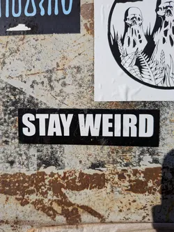 The sticker is a horizontal rectangle with a solid black background. The phrase 'STAY WEIRD' is written in prominent, all-caps, bold white sans-serif font. The letters are clean and blocky. The sticker shows minor signs of wear, with slight scuffs on the white text and some texture visible on the black background, suggesting it's adhered to a rough, possibly rusty, surface.