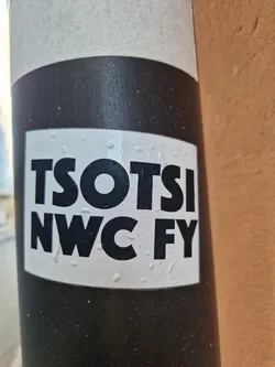 Street sticker A black and white sticker with the text "TSOTSI NWC FY" in a bold, sans-serif font. The sticker is affixed to a dark gray pole.  The sticker shows some minor water droplets.