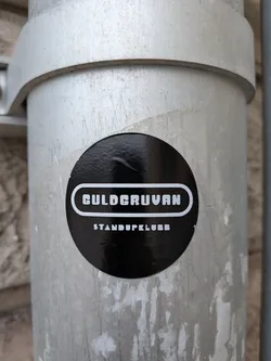 Street sticker The sticker is circular and predominantly black with white text. The text is arranged in two lines. The top line features the word 'GULDCURVAN' in a stylized, bold, sans-serif font with a white outline. Below this, in smaller, all-caps sans-serif font, is 'STANDUPKLUBB'. The sticker has a glossy finish and is slightly curved, adhering to a metallic cylindrical surface.