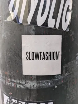 Street sticker The image features a square sticker attached to a dark, cylindrical pole. The sticker is predominantly white with the text "SLOWFASHION" printed in bold, black letters. The word is presented in all caps, and there is a trademark symbol to the right of the word. The pole appears to be made of metal and has other stickers.