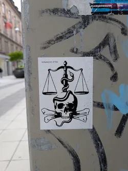 Street sticker The sticker is a black and white graphic design on a white rectangular background. It depicts a central image of a human skull with two crossbones positioned beneath it. Above the skull, a classic set of scales of justice is shown, with two empty pans hanging from a central beam. A snake is coiled around the vertical pillar of the scales, with its head positioned near one of the scale pans. The entire design is rendered in bold black lines and solid black fills, creating a high-contrast, symbolic image. Small water droplets are visible on the surface of the sticker, indicating it's outdoors. In the top-left corner, the text 'IGNACIO.TTD' is printed.
