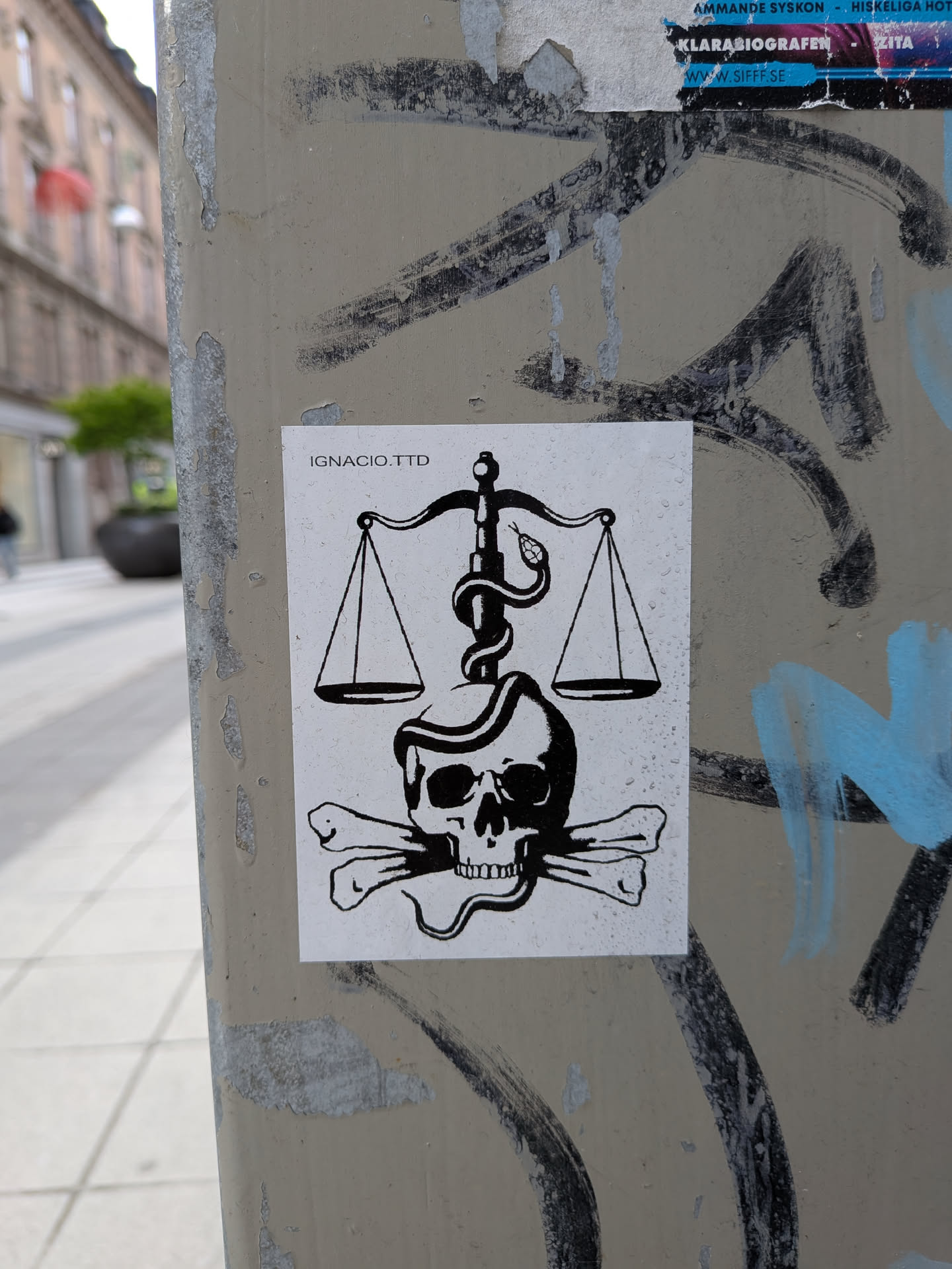 The sticker is a black and white graphic design on a white rectangular background. It depicts a central image of a human skull with two crossbones positioned beneath it. Above the skull, a classic set of scales of justice is shown, with two empty pans hanging from a central beam. A snake is coiled around the vertical pillar of the scales, with its head positioned near one of the scale pans. The entire design is rendered in bold black lines and solid black fills, creating a high-contrast, symbolic image. Small water droplets are visible on the surface of the sticker, indicating it's outdoors. In the top-left corner, the text 'IGNACIO.TTD' is printed.
