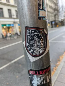 Street sticker A black and white sticker on a metal pole depicts a wolf's head within a shield-shaped emblem.  Three upward-pointing trident-like symbols sit above the wolf. The style is dark and possibly related to occult or metal music imagery. 