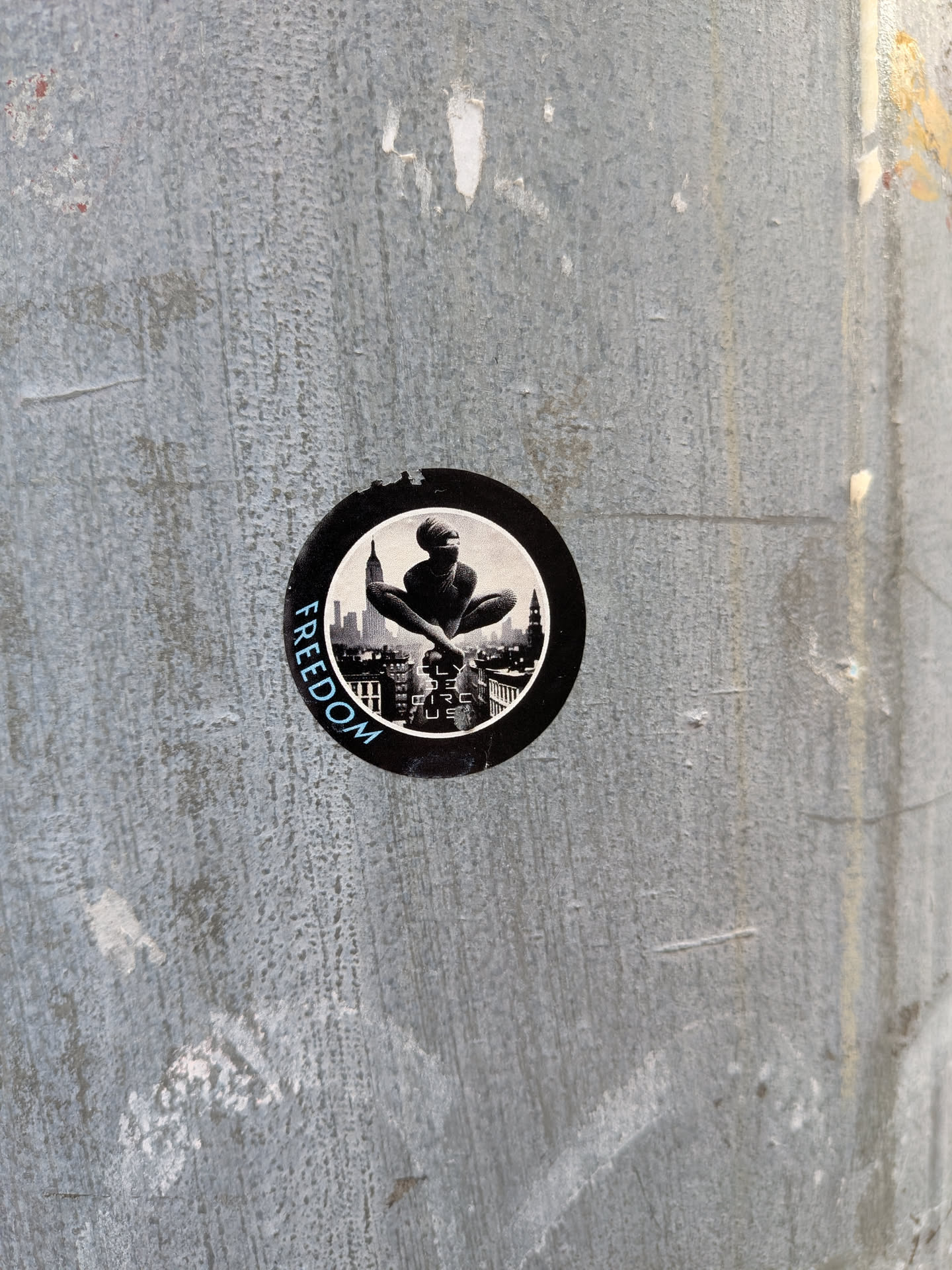 A circular sticker with a black border. The central image is a black and white depiction of a person in a meditative pose, sitting cross-legged with their arms resting on their knees. Behind the figure is a detailed cityscape with tall buildings and a prominent skyscraper resembling the Empire State Building. The word 'FREEDOM' is written in a light blue, stylized font in an arc along the left side of the sticker's border. Below the figure, within the cityscape, there appears to be some very small, illegible text.