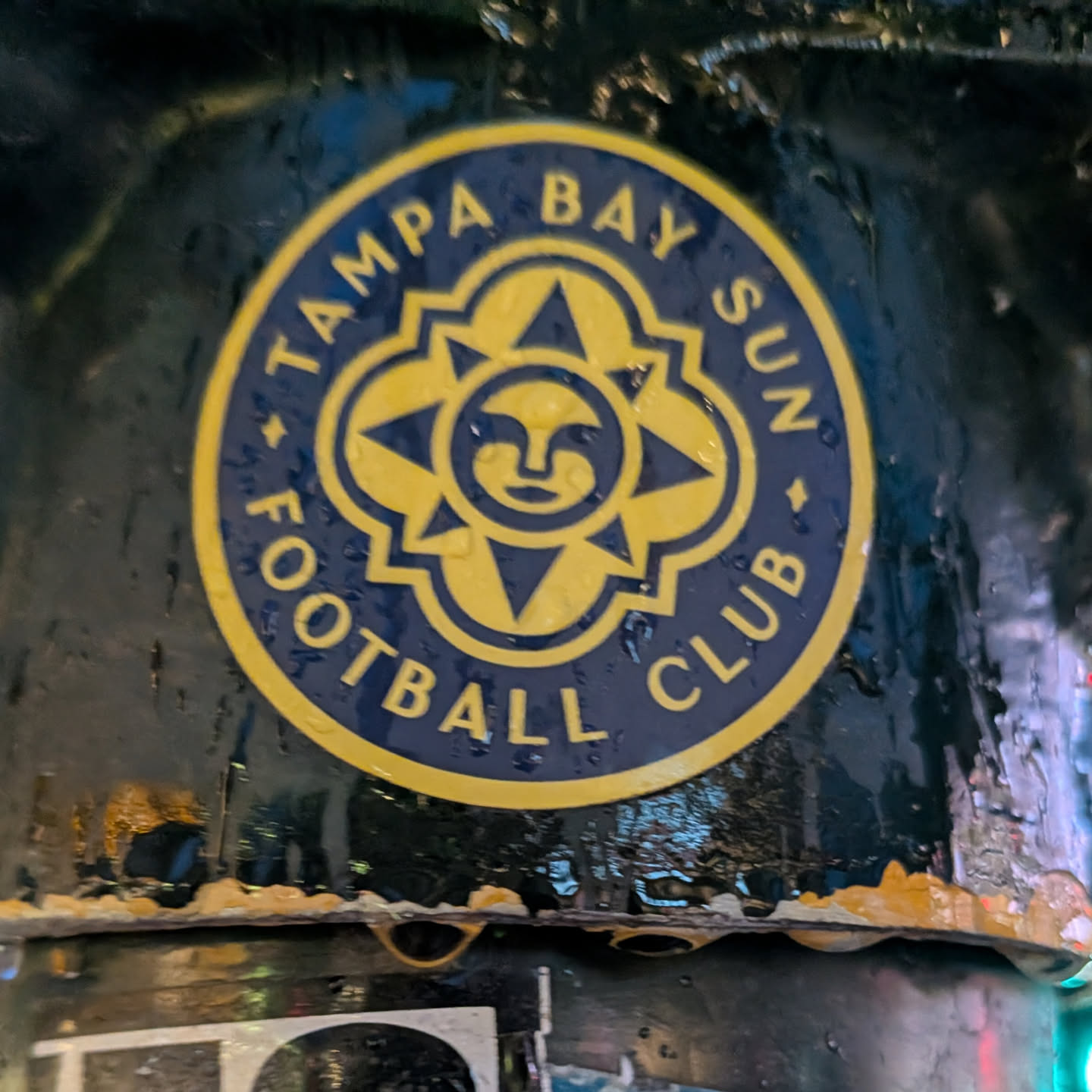The sticker is circular with a dark blue background and a yellow border. In the center, there is a stylized yellow sun with a face. The sun has four points extending outwards, with smaller triangular shapes within each point. The text 'TAMPA BAY SUN FOOTBALL CLUB' is written in yellow capital letters around the circumference of the sticker, separated by small yellow dots.