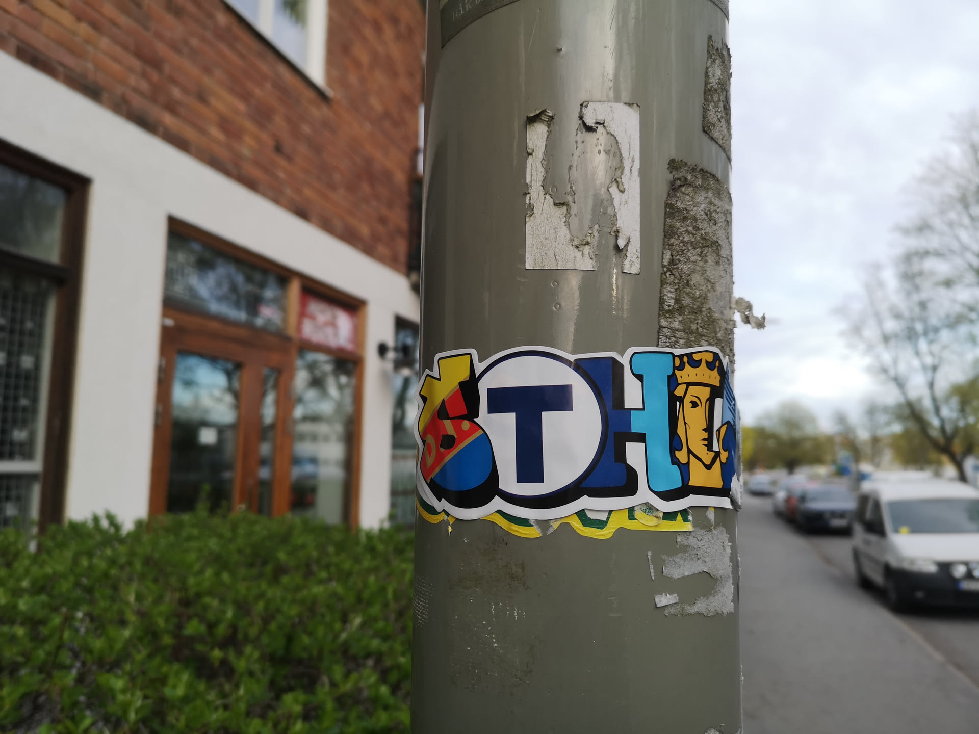The sticker is a multi-part, colorful design with a thick black outline and a white border, adhered to a grey pole. On the far left, there's a stylized emblem or shield in red, yellow, and blue, possibly representing an 'S'. In the center, a prominent dark blue 'T' is enclosed within a white circle, strongly resembling a public transit or subway logo. To the right, a light blue block features a yellow crowned head, possibly a king or queen, facing right, with a light brown face. Below the main design, there's a wavy line in yellow and green. The overall design appears to spell 'STH' using these distinct graphic elements.