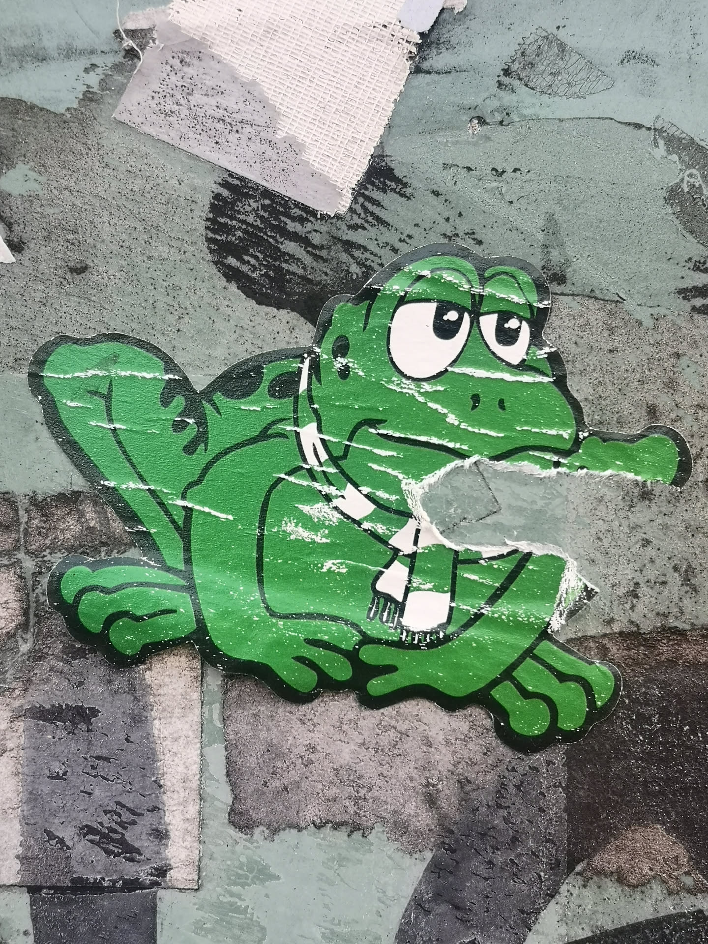 A cartoon-style sticker depicting a bright green frog with a somewhat grumpy or sad expression. The frog has large white eyes with black pupils and thick black outlines. It is wearing a white scarf with black stripes around its neck. The sticker shows significant wear and tear, with numerous white scuff marks and areas where the print has peeled, particularly across its body and around its mouth. The frog is positioned in a crouching or sitting pose, with its limbs visible. It is affixed to a textured, multi-colored surface that appears to be a wall or panel, showing shades of grey, muted green, and black, with remnants of other materials.