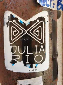 Street sticker A white sticker with a black M-shaped design containing spiraled triangle shapes, above the name "JULIA RIO" also in black. The sticker has visible wear and tear, especially around the edges. It is stuck to a rusted metal pole.