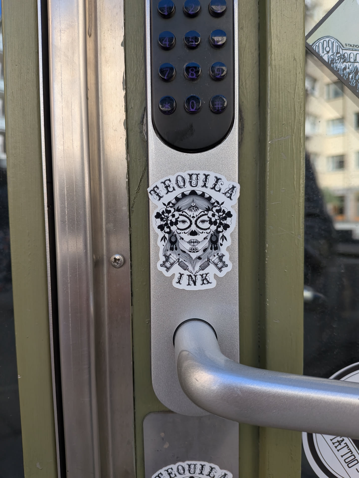 The sticker showcases a detailed, black and white sugar skull (Calavera) design with a clean white border. The skull features intricate patterns around its eyes, nose, and mouth, characteristic of Day of the Dead artistry. It is adorned with a headpiece of large, dark flowers and leaves on both sides. Below the skull, two tattoo machines are crossed, their needles pointing downwards. The text 'TEQUILA' is arched above the skull, and 'INK' is positioned below it, both in a decorative, gothic-style font.