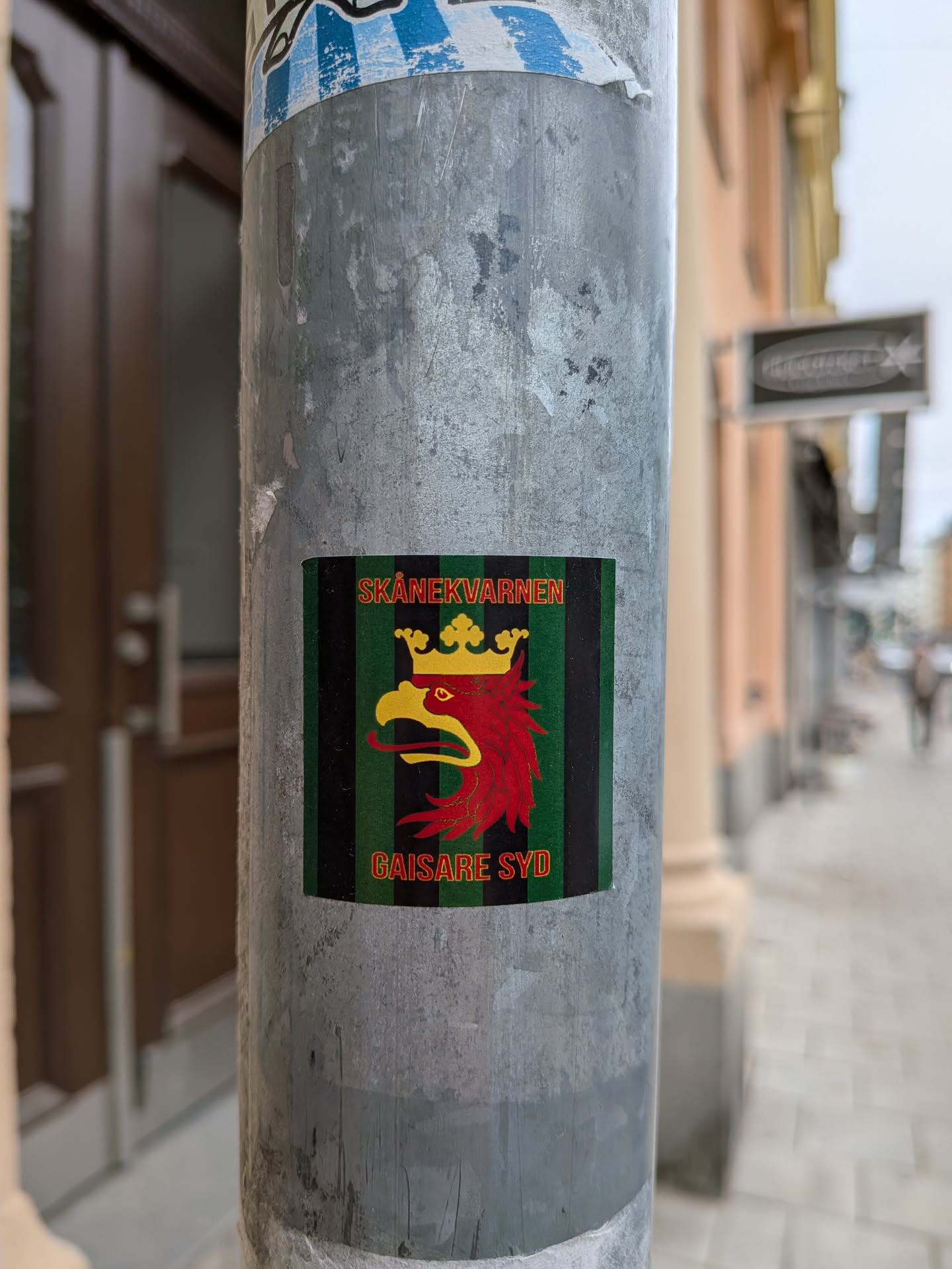 A rectangular sticker featuring a stylized red griffin head with a yellow beak and tongue, adorned with a golden crown. The griffin is set against a background of alternating vertical stripes in dark green and black. Above the griffin, in yellow capital letters, is the text 'SKÅNEKVARNEN', and below it, also in yellow capital letters, is 'GAISARE SYD'. The overall design is bold and heraldic, suggesting regional pride and affiliation.