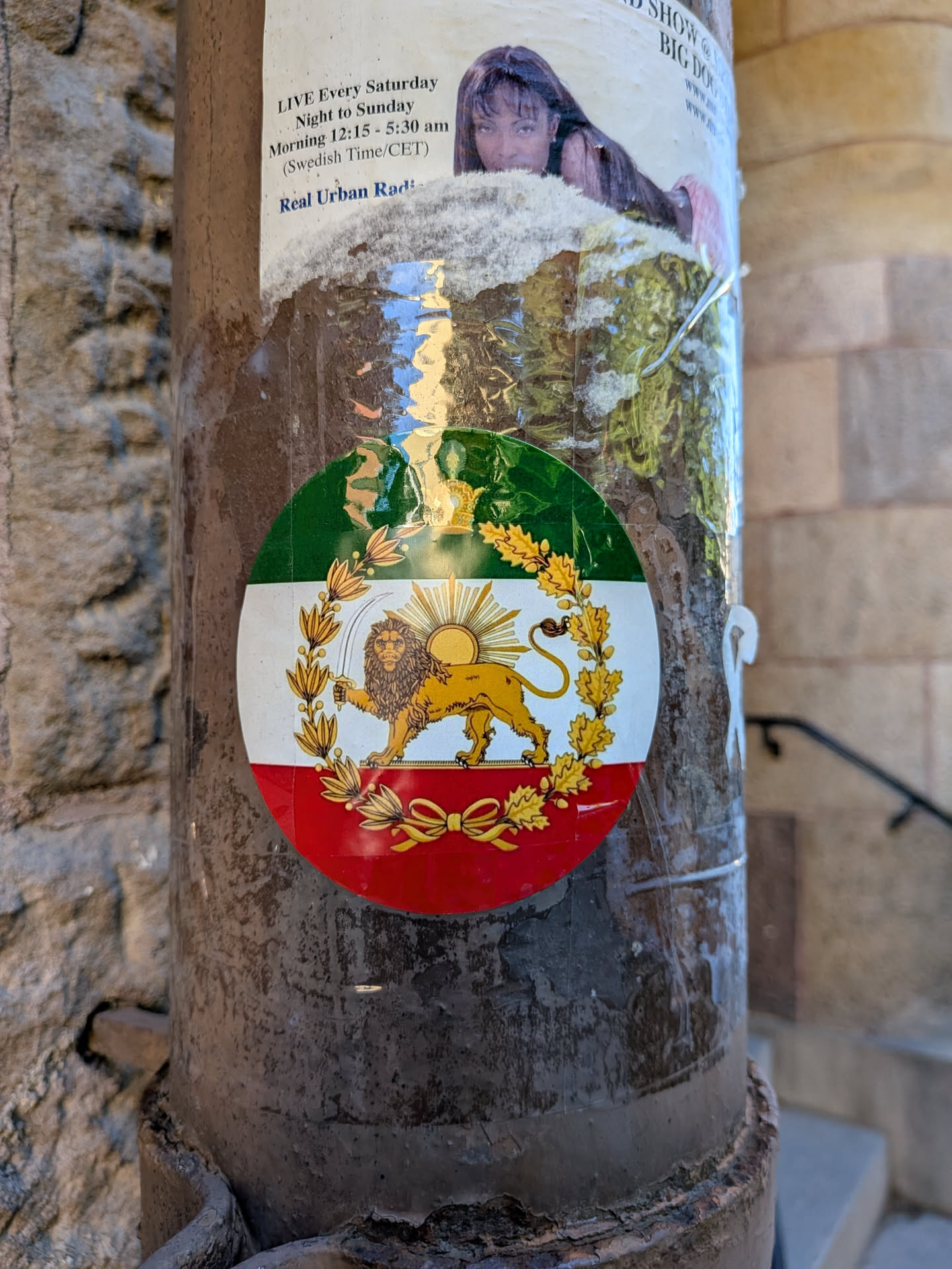A round sticker displaying the Iranian Lion and Sun symbol over a green, white, and red tricolor background. The golden lion holds a sword with a rising sun behind it, enclosed in a golden wreath.