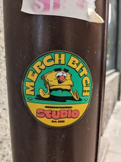 Street sticker A round sticker featuring a cartoon illustration of a piece of toasted bread with a face. The sticker is predominantly teal green with yellow and orange accents. Text on the sticker reads: 'MERCH BITCH' in a circular design and 'SCREENPRINTING STUDIO Est. 2015' in smaller text below the character. The sticker is affixed to a brown pole.