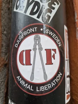 Street sticker A round sticker with a black background and red and white inner circle.  The inner circle shows a pair of bolt cutters and the letters "D" and "F" in red. The text "Djurfront • Sweden" appears in a circular arc above the image, and "Animal Liberation" appears below. 