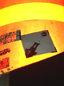 The central sticker is a rectangular print, predominantly grayscale, depicting a tall, dark bell tower or steeple. The tower features an open belfry with a bell visible inside, and a pointed roof topped with a weather vane. Several small, dark bird silhouettes are shown flying around the upper part of the tower against a lighter, muted sky background. Above the tower, a small, dark, irregular shape resembling a cloud contains faint white text. The sticker has a thin, dark border. The warm orange and yellow light from the background illuminates the sticker, giving its edges and the lighter parts of the image a warm hue, despite the original print likely being monochromatic.