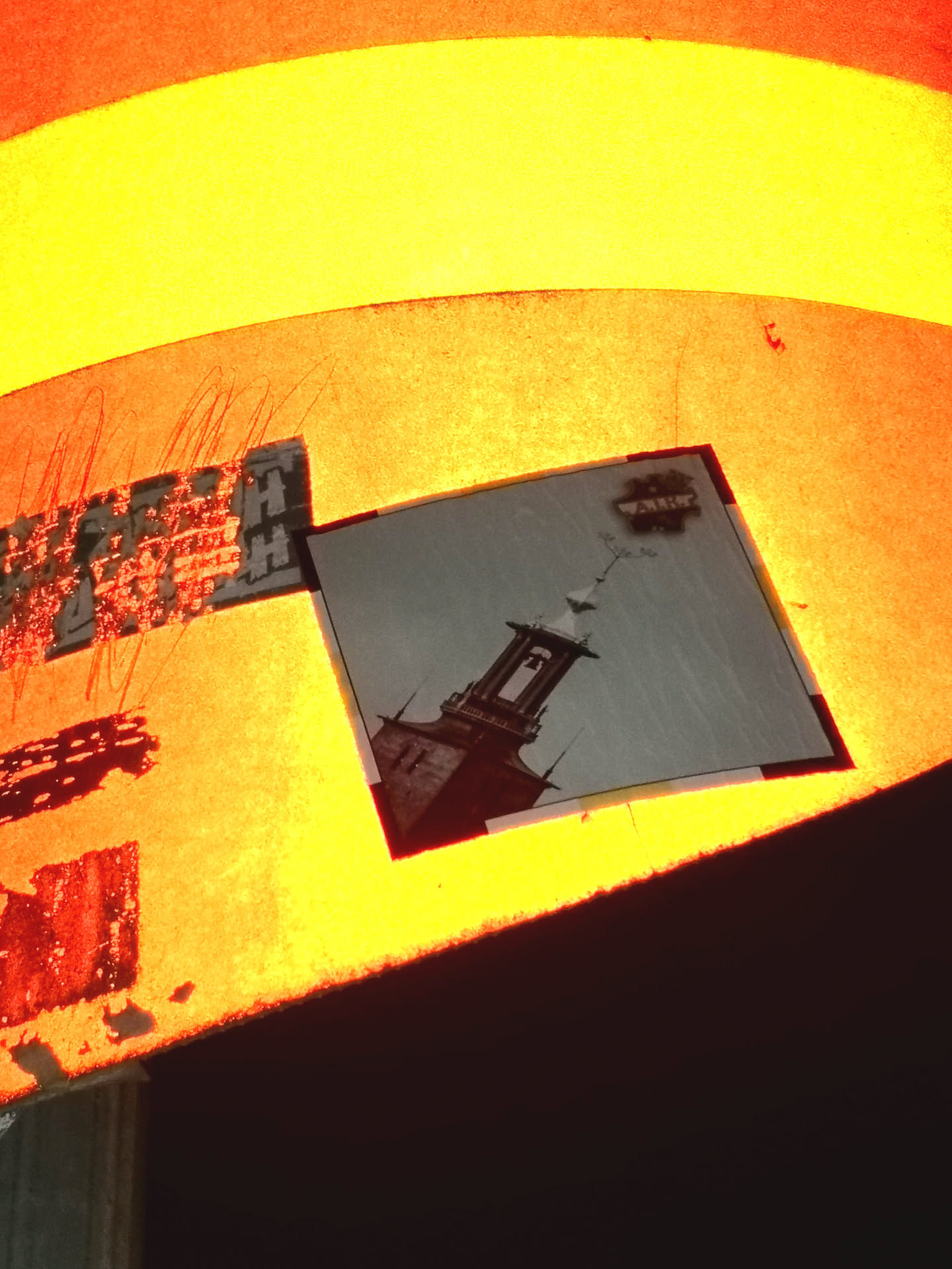 The central sticker is a rectangular print, predominantly grayscale, depicting a tall, dark bell tower or steeple. The tower features an open belfry with a bell visible inside, and a pointed roof topped with a weather vane. Several small, dark bird silhouettes are shown flying around the upper part of the tower against a lighter, muted sky background. Above the tower, a small, dark, irregular shape resembling a cloud contains faint white text. The sticker has a thin, dark border. The warm orange and yellow light from the background illuminates the sticker, giving its edges and the lighter parts of the image a warm hue, despite the original print likely being monochromatic.