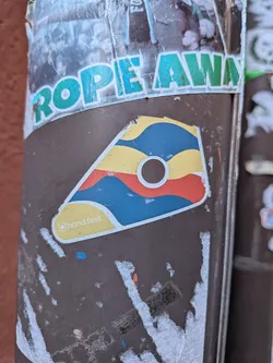 Street sticker The sticker is an irregular, somewhat triangular or trapezoidal shape with rounded corners, resembling a stylized guitar pick or shield. It features a bold, abstract design composed of wavy, horizontal bands of color: yellow at the top, followed by blue, then red, and another blue band at the bottom. A prominent white circle with a dark brown center is located in the upper-middle section of the sticker. The entire design is outlined in white. In the bottom-left corner, within a yellow section, is the text '@hand.fest'. The colors are bright and distinct, giving it a modern, graphic appearance.