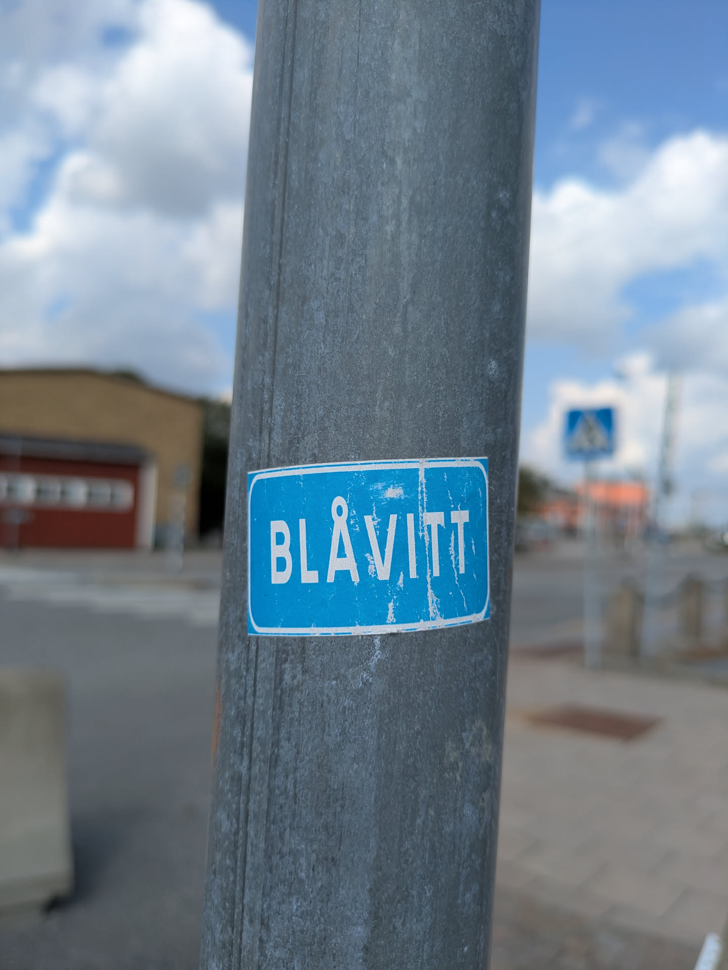 A rectangular sticker with rounded corners featuring a light blue background and the word 'BLÅVITT' in bold, uppercase white letters. The sticker shows signs of wear, including faded colors, chipped or flaking white text, and slightly curled edges, indicating exposure to the elements. It is adhered to a textured, grey metal pole.