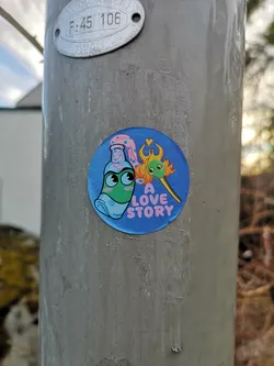 Street sticker The sticker is circular with a vibrant blue background. On the left, a light green bottle with large, expressive eyes and a light blue mask-like feature is depicted. It wears a pink cap adorned with small white flowers or dots, from which a pink, wavy, cloud-like element extends upwards. To the right, a yellow matchstick with a small green head and eyes is shown, its tip engulfed in orange and yellow flames. Above the matchstick's flame, a small red heart is visible. Below the matchstick, the words "A LOVE STORY" are written in pink, sans-serif font. The overall style is cartoonish and cheerful.