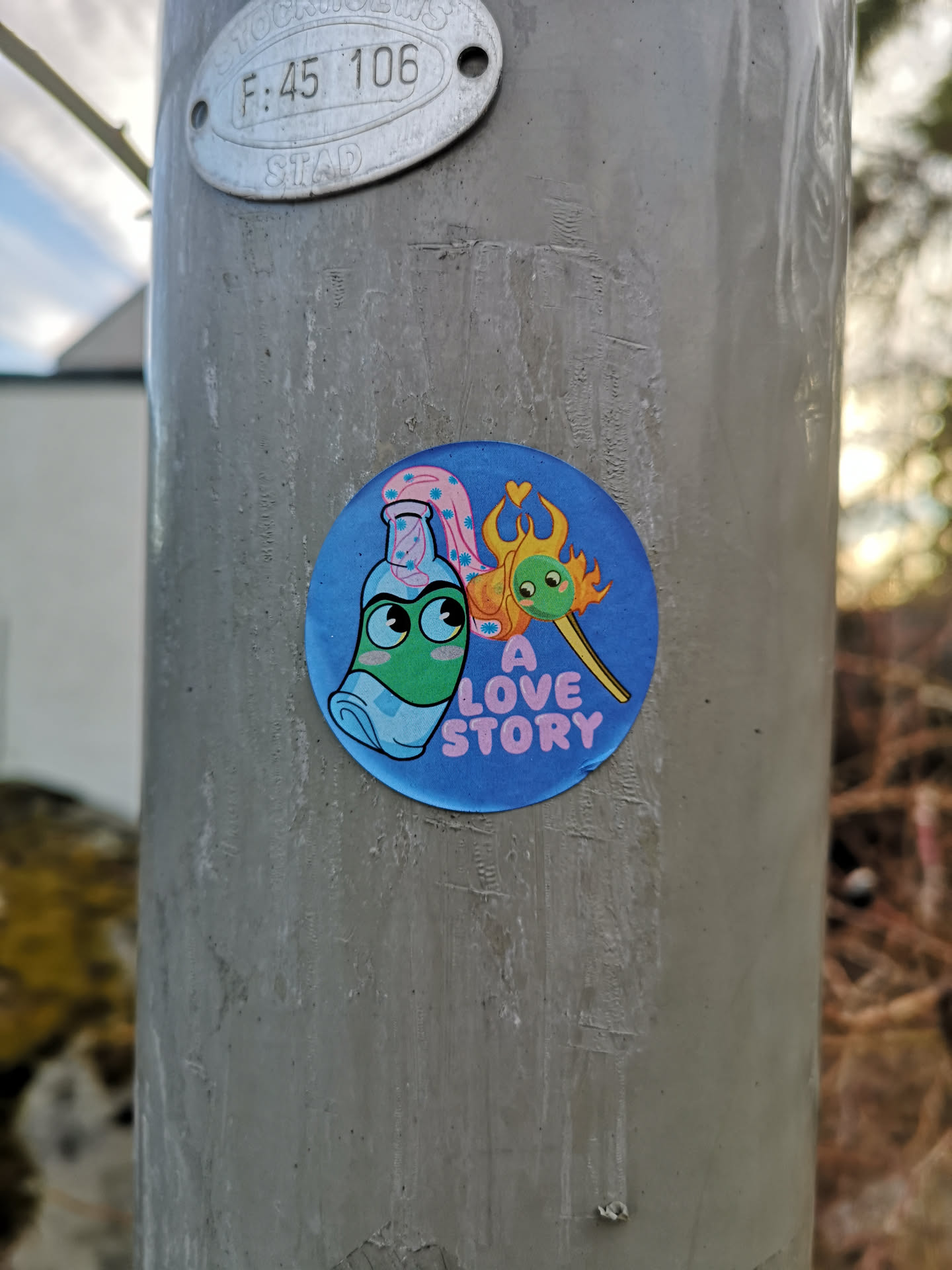 The sticker is circular with a vibrant blue background. On the left, a light green bottle with large, expressive eyes and a light blue mask-like feature is depicted. It wears a pink cap adorned with small white flowers or dots, from which a pink, wavy, cloud-like element extends upwards. To the right, a yellow matchstick with a small green head and eyes is shown, its tip engulfed in orange and yellow flames. Above the matchstick's flame, a small red heart is visible. Below the matchstick, the words "A LOVE STORY" are written in pink, sans-serif font. The overall style is cartoonish and cheerful.