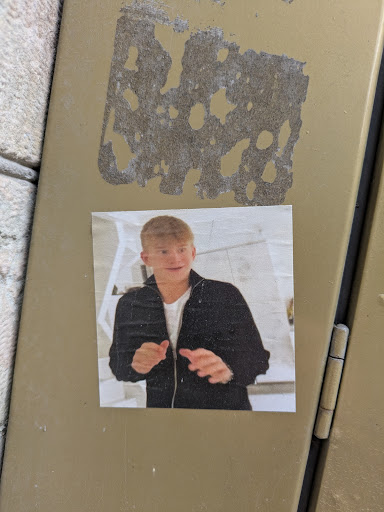 The sticker features a photo of a fair-skinned person with short blond hair wearing a dark jacket and white shirt. The background appears to be a modern interior with white walls and geometric elements. The sticker is adhered to a somewhat worn metal surface.