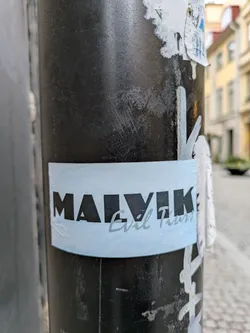 A light blue sticker with the text "MALVIK" in bold black letters and "Evil Tours" written in a smaller, italicized font below it. The sticker is affixed to a dark gray pole.