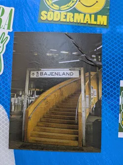 Street sticker The sticker displays a photograph of a subway or metro station entrance. A prominent, wide, curved staircase with yellow-orange steps and walls dominates the center, leading upwards. A silver handrail runs along the curve of the stairs. Above the staircase, a white rectangular sign with black text and an upward arrow clearly indicates "BAJENLAND". The ceiling is dark with visible fluorescent light fixtures, casting a somewhat dim and industrial light on the scene. The walls surrounding the stairs appear to have some graffiti, adding to a gritty, urban aesthetic. The overall image captures a moment within a bustling public transport environment.