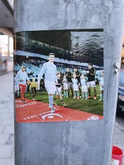 Street sticker A sticker featuring a photo of a male footballer in a light blue uniform, walking on a red carpet. He is surrounded by a line of young boys, also in light blue uniforms, who appear to be part of a football team.  The background shows a stadium. The sticker has a logo at the bottom that resembles a crescent moon.