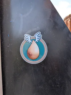 The sticker features a central, inverted teardrop or egg-shaped form in a gradient of light brown/skin tone, with subtle shading. This shape is set within a circular design, which has a wide teal-blue inner ring, bordered by a thin light grey line, and then a white outer edge. Perched atop the central teardrop, extending slightly beyond the inner teal ring, are two stylized white wings with light blue outlines. The overall aesthetic is clean, illustrative, and cartoon-like.