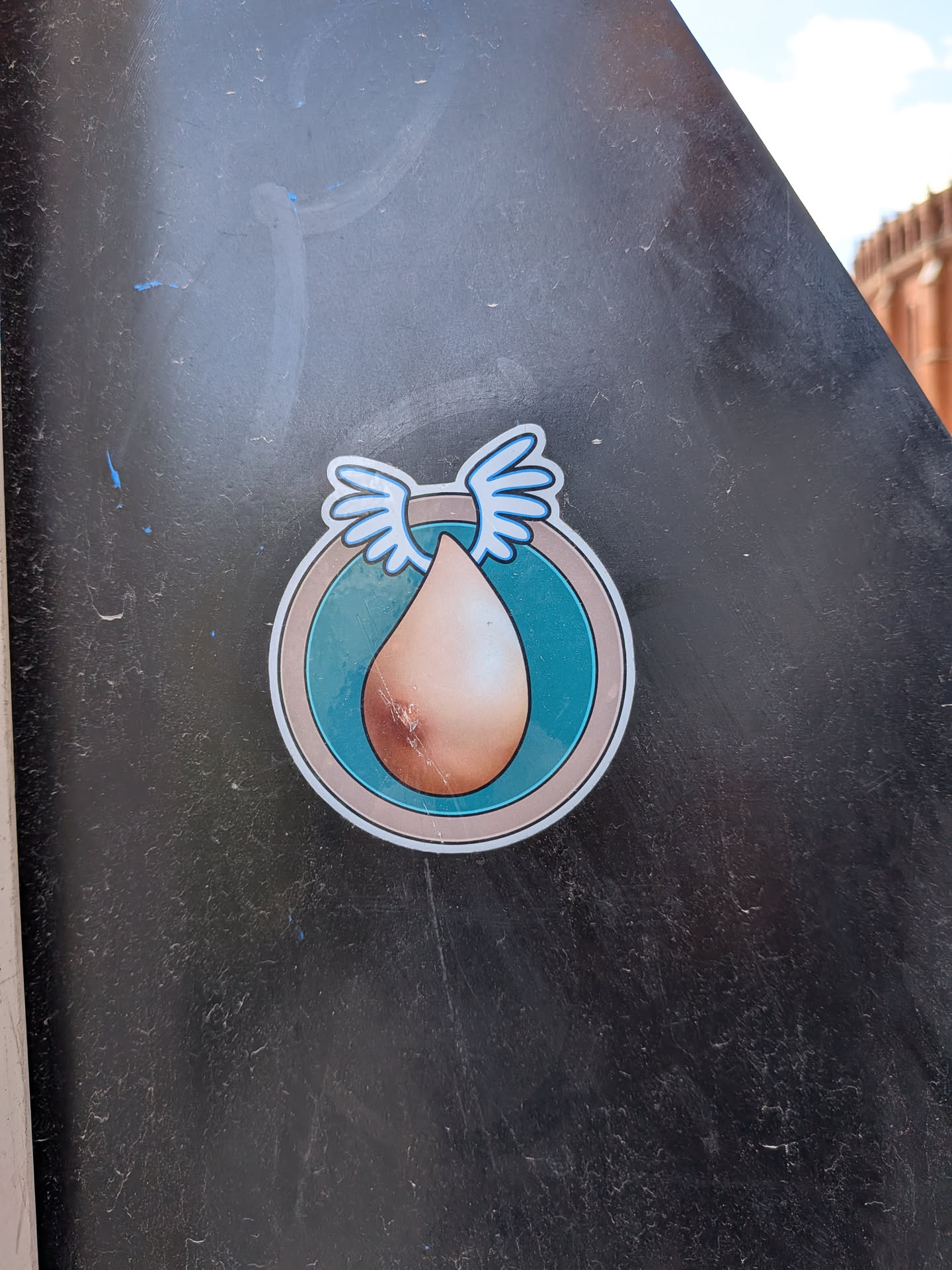 The sticker features a central, inverted teardrop or egg-shaped form in a gradient of light brown/skin tone, with subtle shading. This shape is set within a circular design, which has a wide teal-blue inner ring, bordered by a thin light grey line, and then a white outer edge. Perched atop the central teardrop, extending slightly beyond the inner teal ring, are two stylized white wings with light blue outlines. The overall aesthetic is clean, illustrative, and cartoon-like.